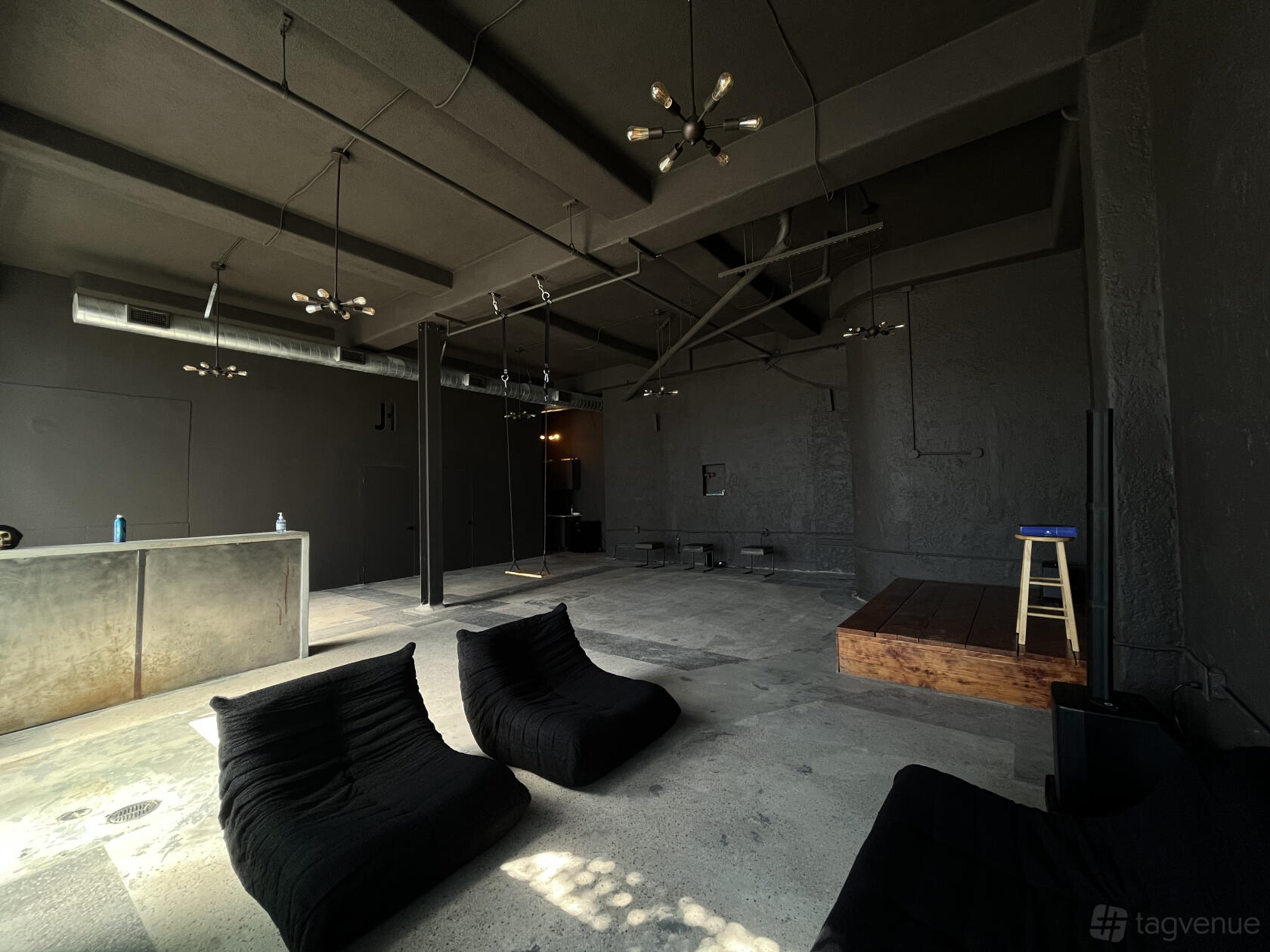 An event venue with black lounge chairs, exposed ductwork, and a small wooden stage at The Hype Spot.
