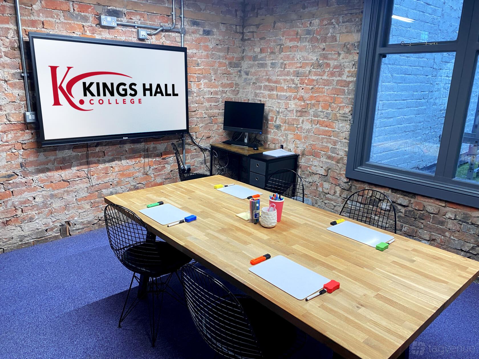 A classroom with exposed brick walls, a wooden table for eight, and a wall-mounted screen at Kings Hall College.
