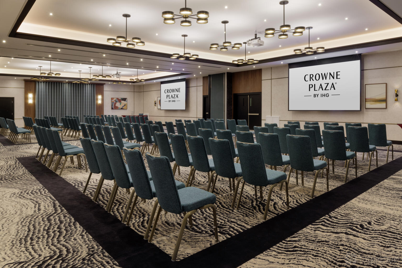 A hotel meeting room with rows of blue chairs facing dual projection screens and patterned carpet at Crowne Plaza London Gatwick.