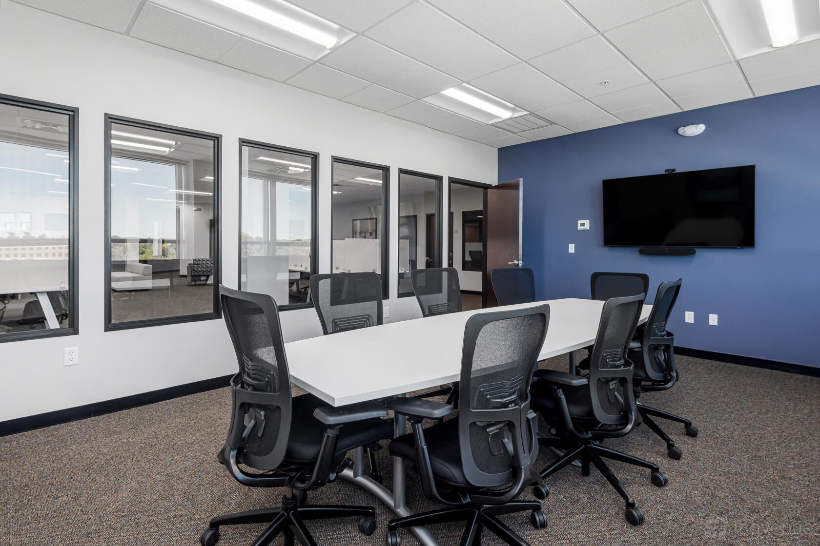 A meeting room with a long white table, black ergonomic chairs, wall-mounted TV, and glass windows at Workspace Beverly.