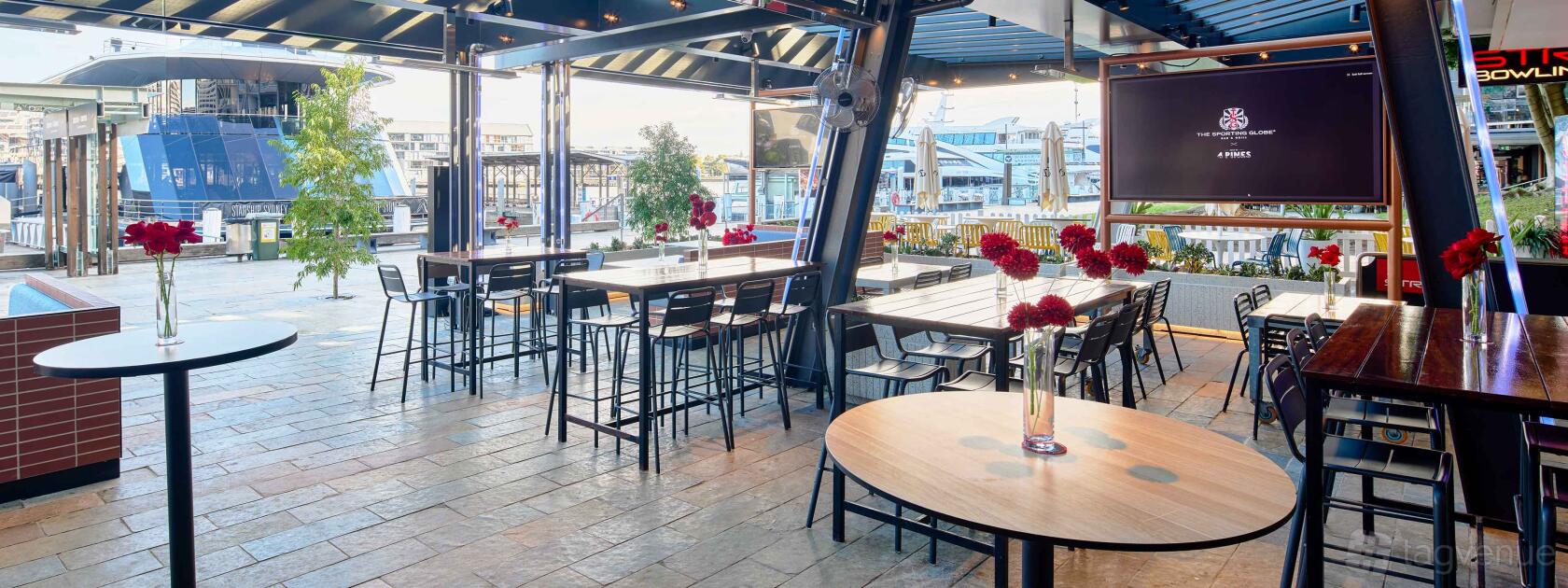 A terrace at a pub with high tables, black chairs, stone flooring, and vases with red flowers at The Sporting Globe King Street Wharf.