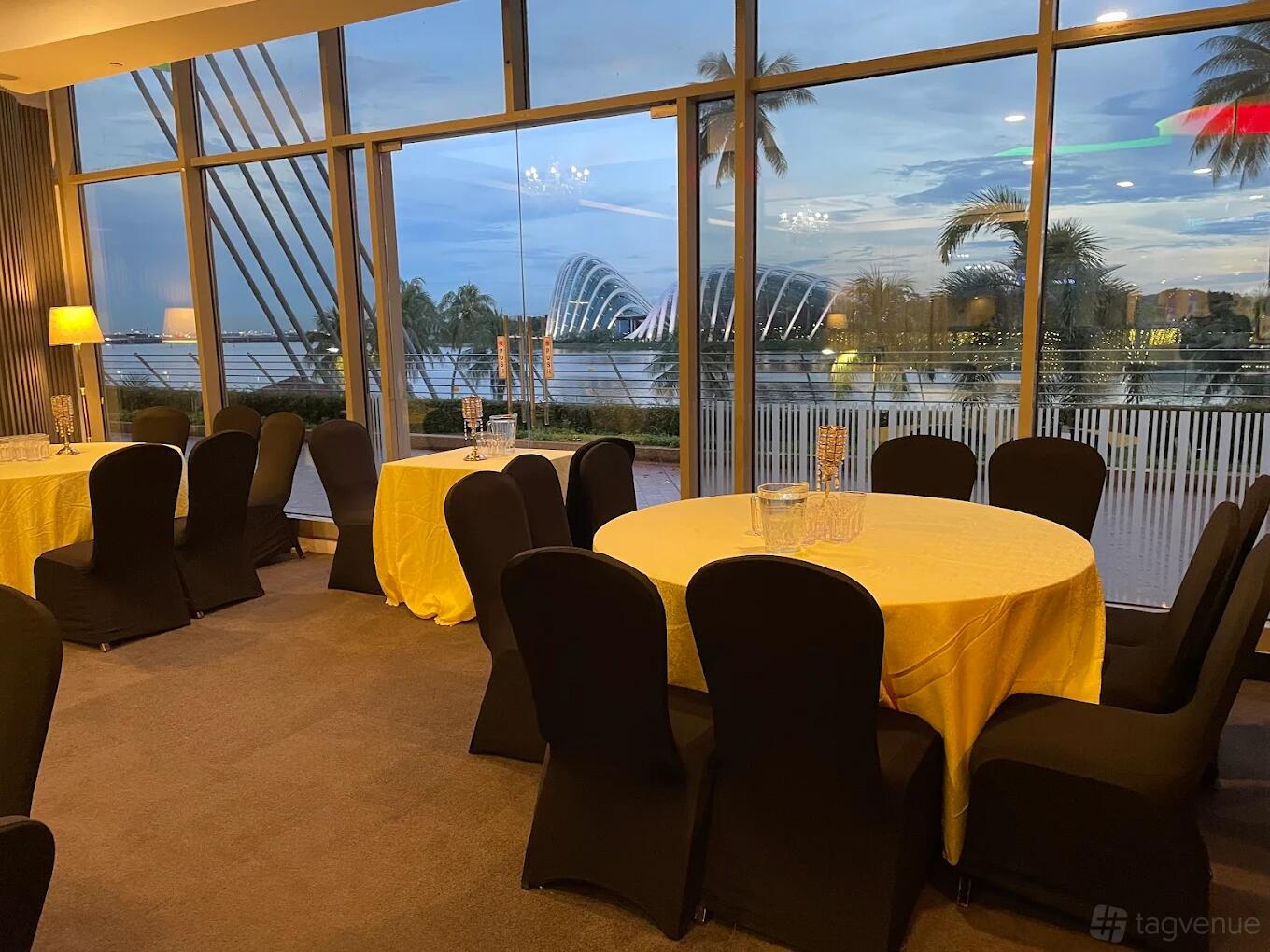 An event space with round tables covered in yellow linens and floor-to-ceiling windows overlooking a waterfront at Red Chillies.