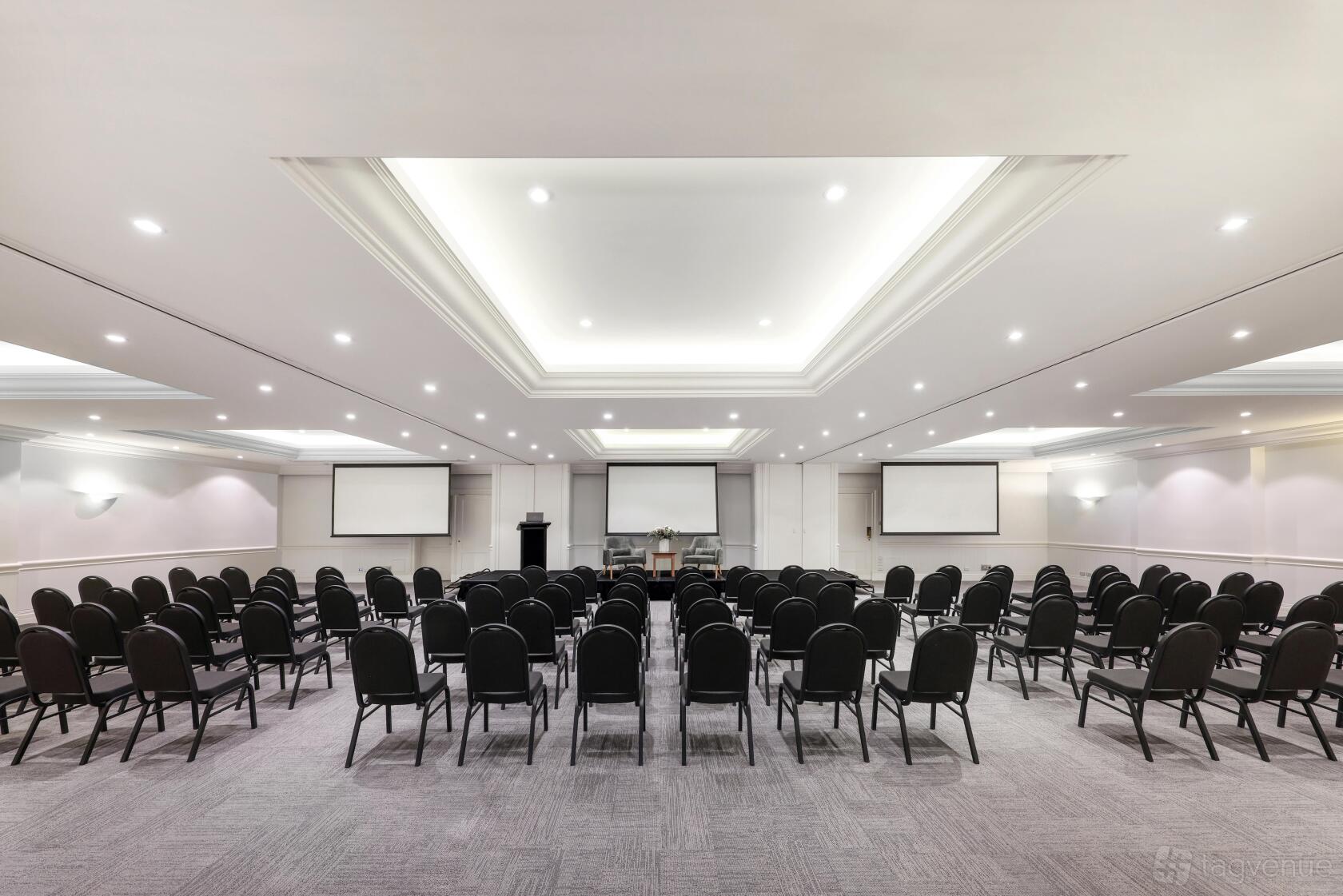 A hotel ballroom with rows of black chairs, grey carpet, and recessed ceiling lighting at Rydges Newcastle.