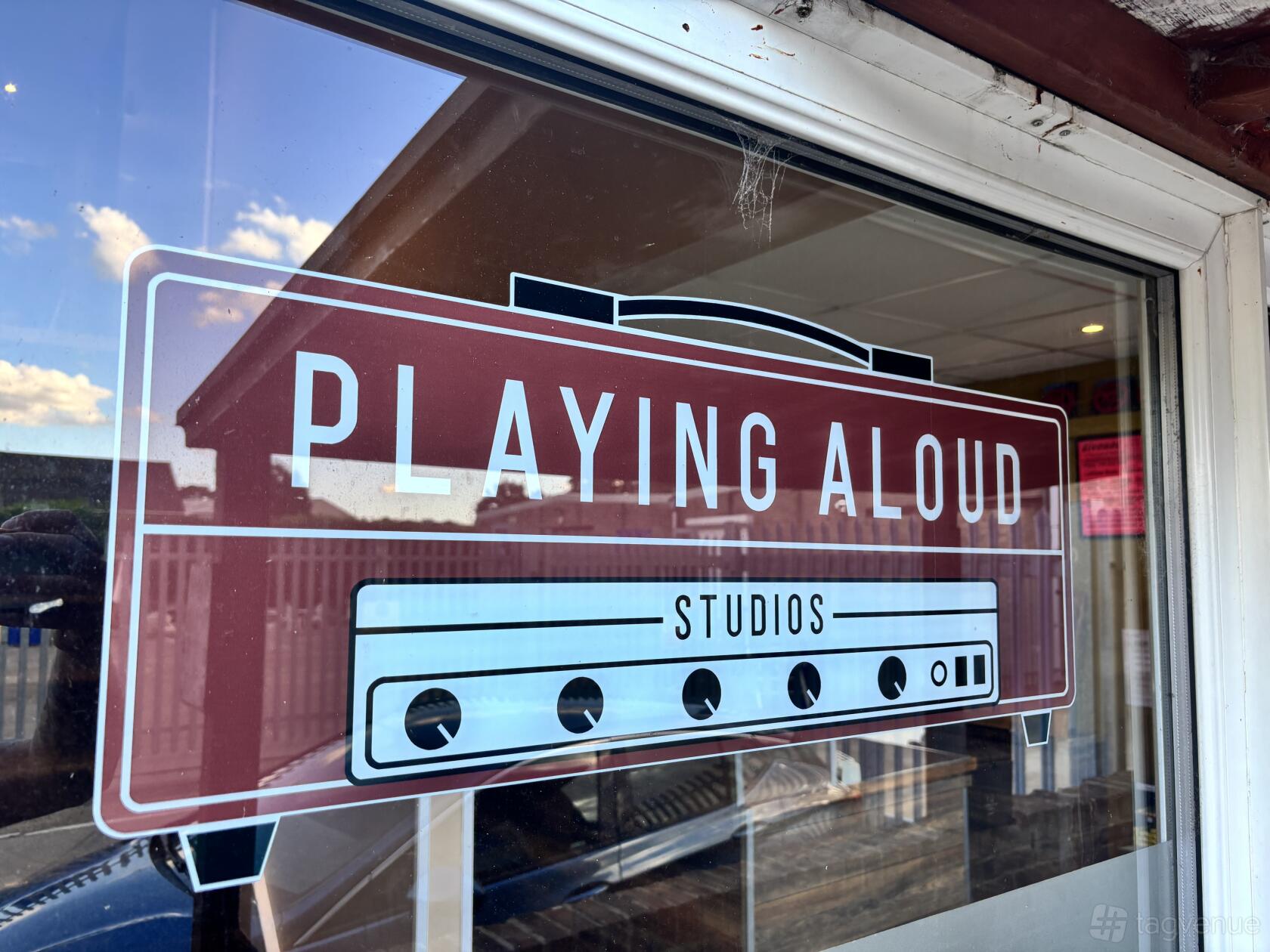 A glass entrance door with the Playing Aloud Studios logo and reflections of a building and sky.