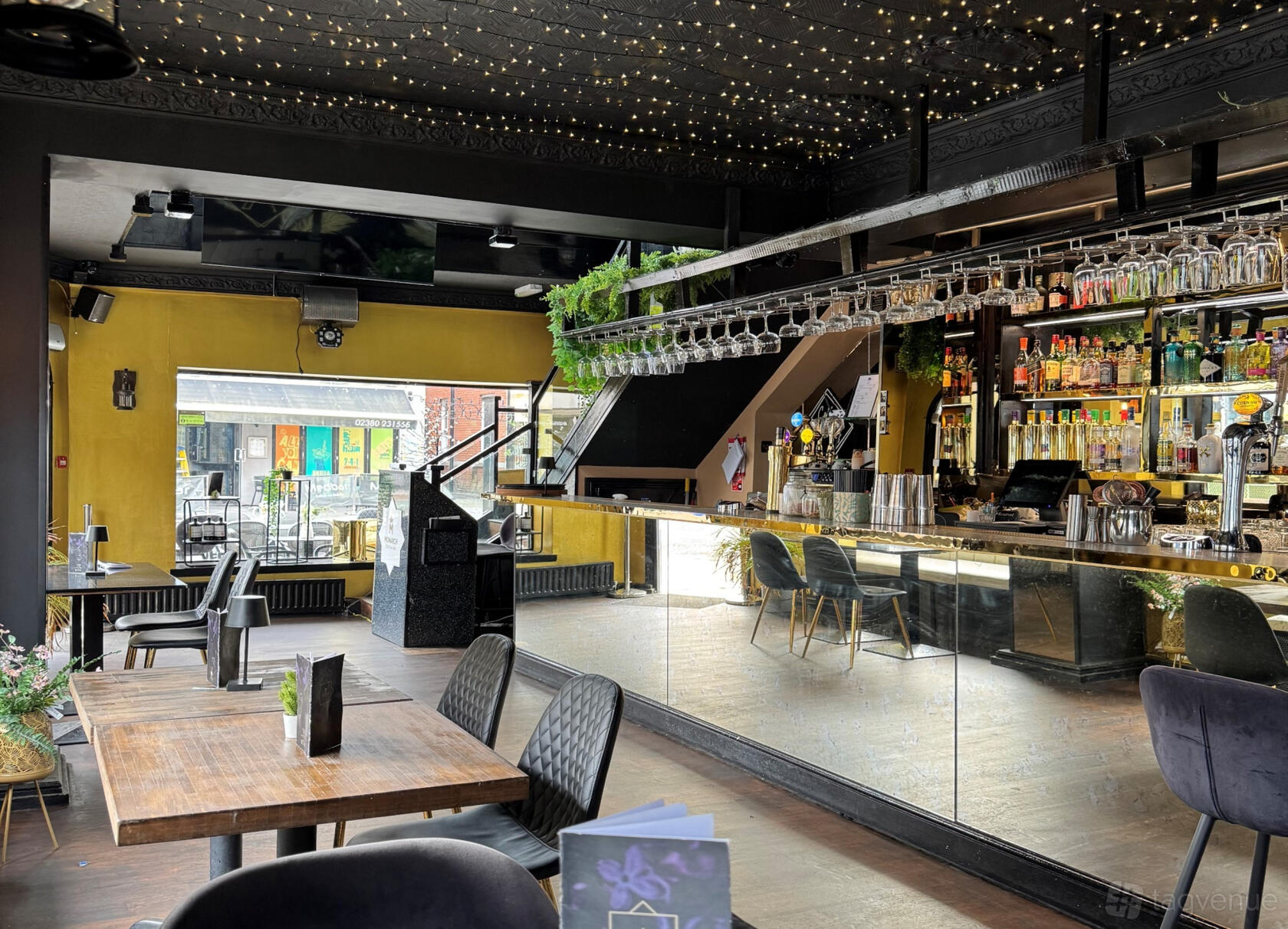 A function room with a mirrored bar, hanging glassware, string lights, and wood tables at Monarch Cocktail Bar.