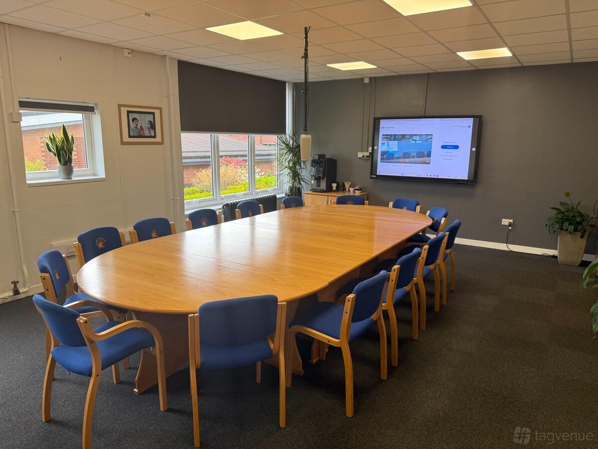 A boardroom with an oval wooden conference table, blue chairs, large windows, and a wall-mounted screen at King Edward VI Five Ways School.