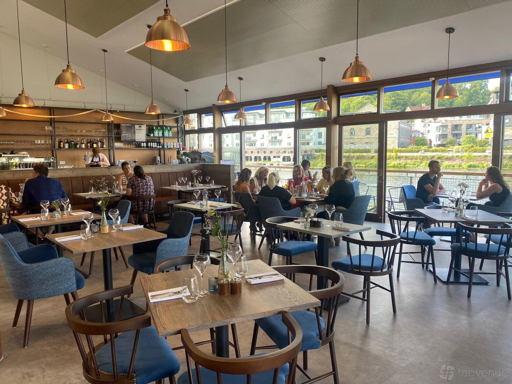 A restaurant with floor-to-ceiling windows, natural light, wooden tables, and blue upholstered chairs at The Board Walk.