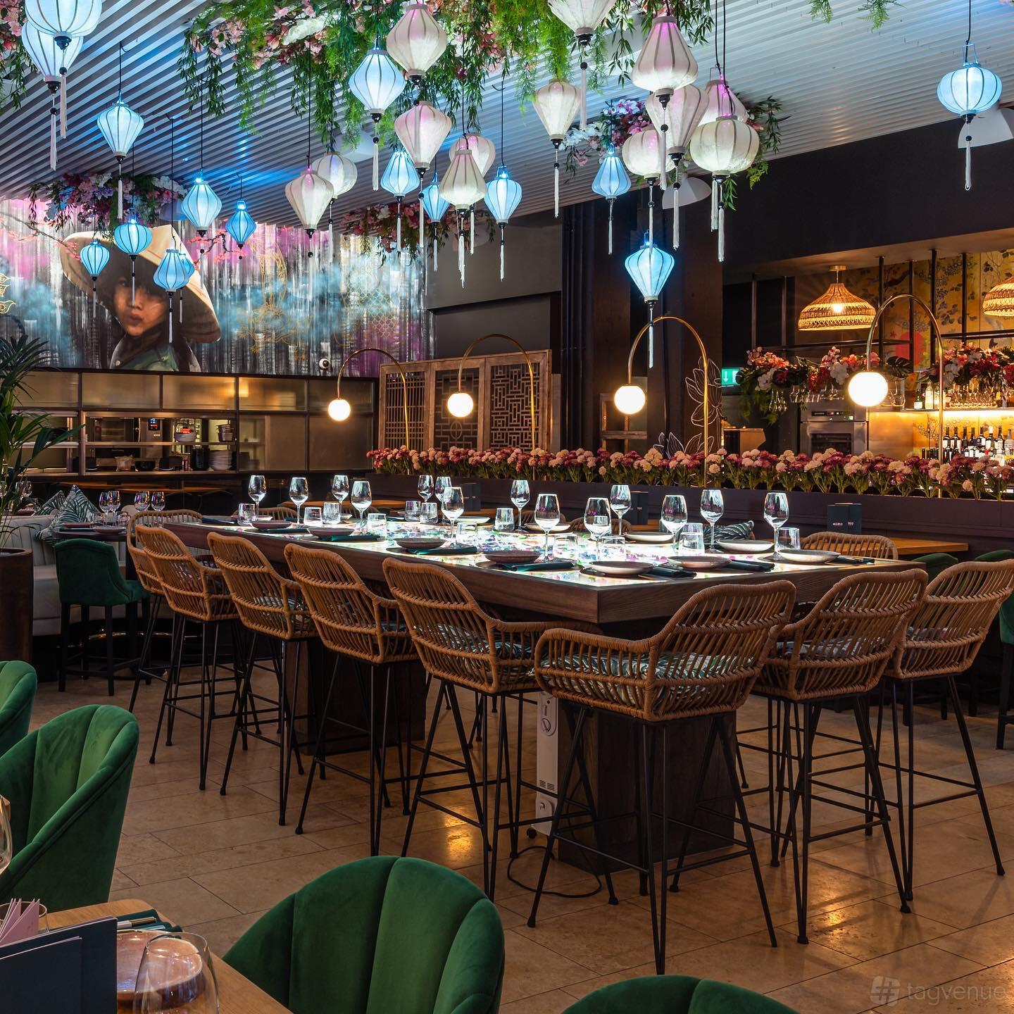 A restaurant with a long wooden table, rattan chairs, floral centerpieces, and blue lanterns at Namii Kitchen & Cocktails.