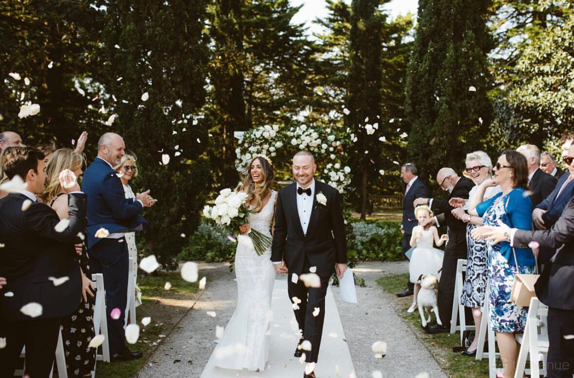 An outdoor ceremony space with a stone pathway, white chairs, and guests surrounded by tall hedges at Como House and Garden.