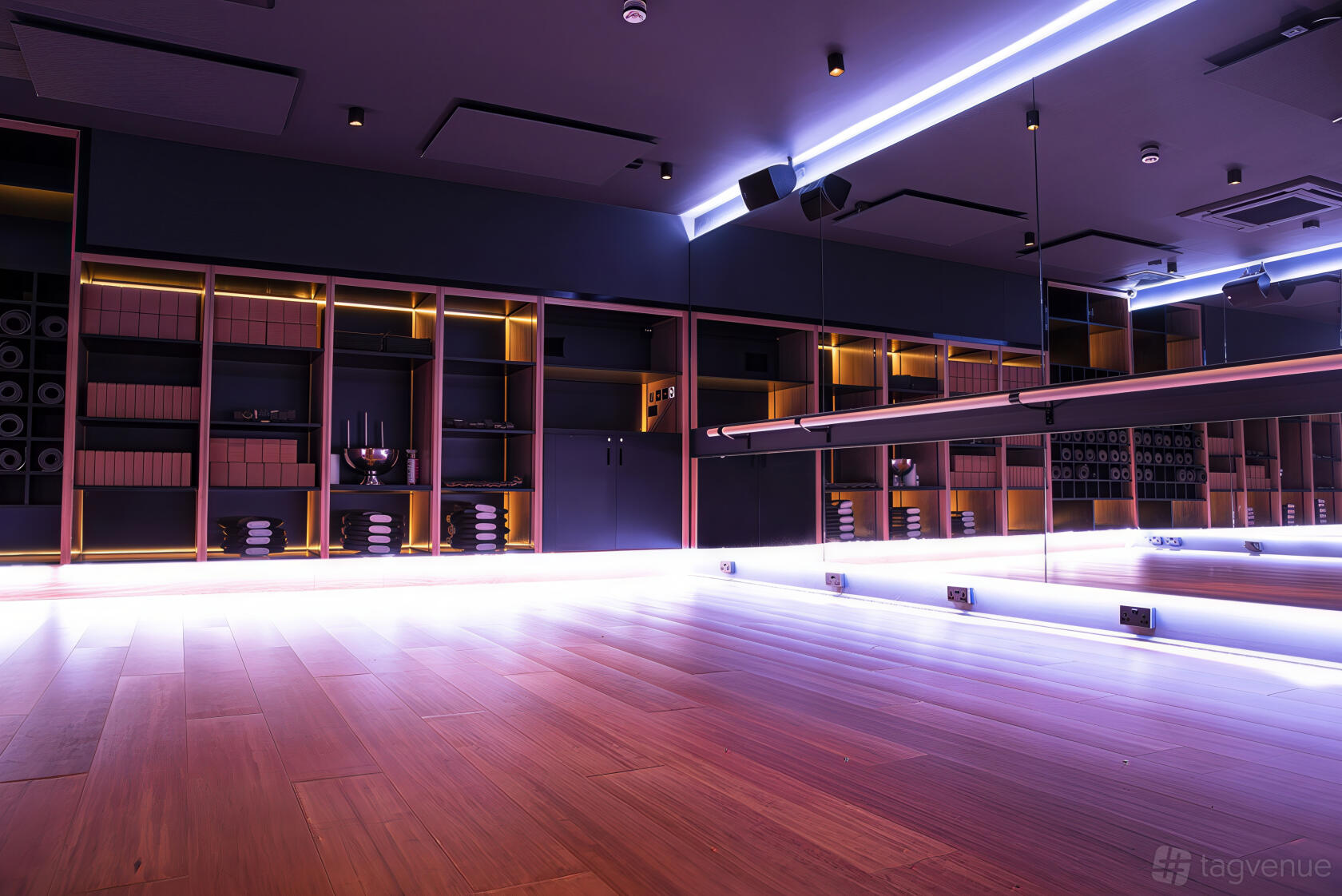 A yoga studio with wood floors, mirrored wall, and shelves with mats and towels at RUMBLE.