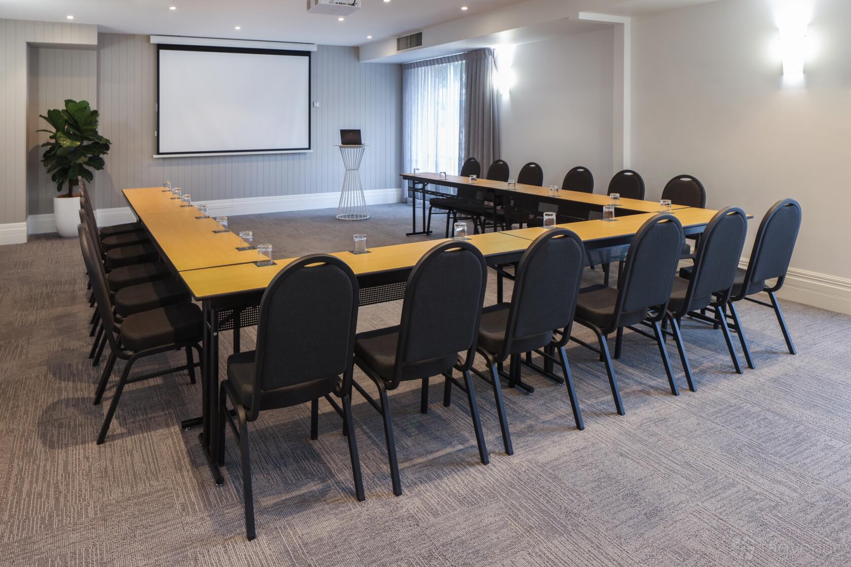 An event space with U-shaped tables, black chairs, a projector screen, and carpeted floors at Rydges Newcastle.