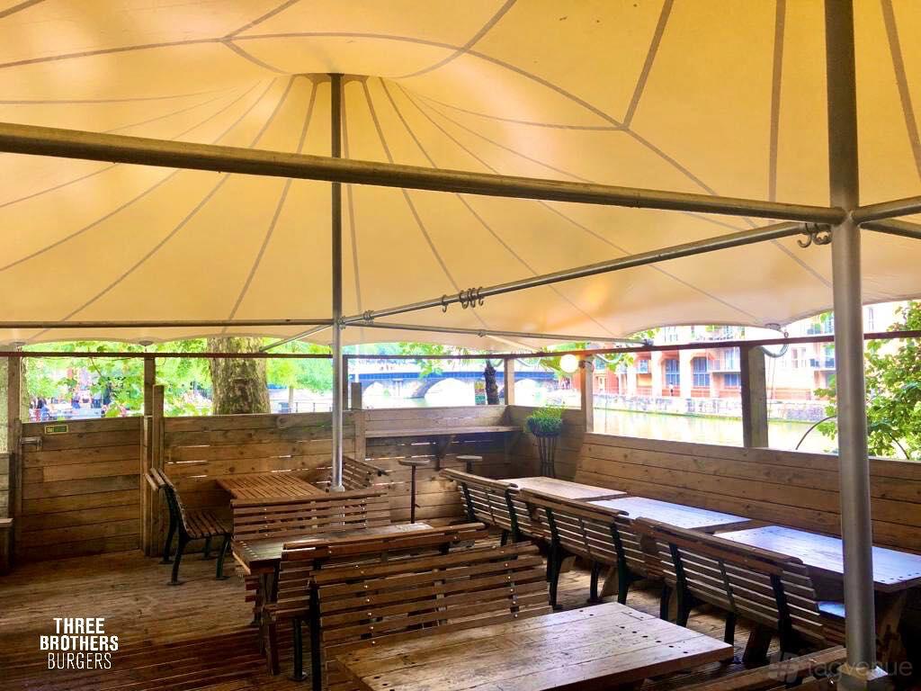 A restaurant terrace with wooden tables and benches under a large canopy at Three Brothers Burgers.