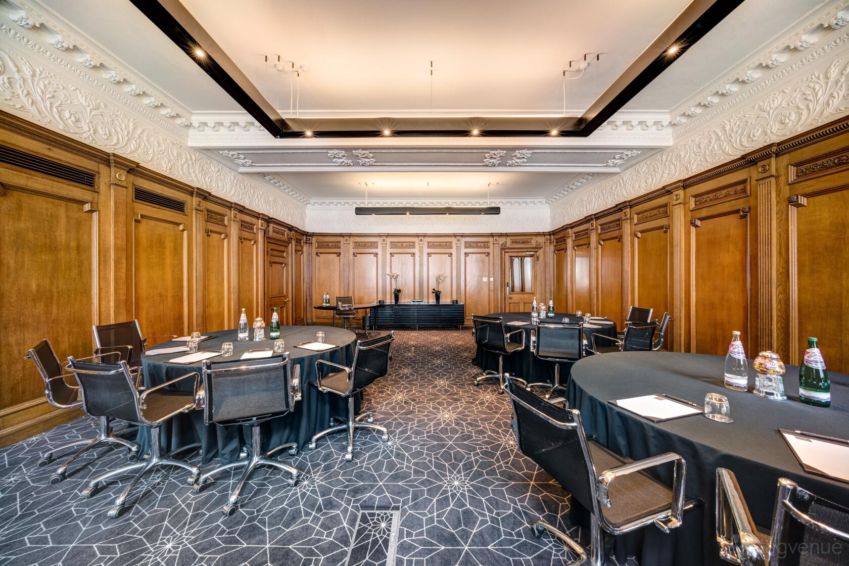 A meeting room with wood-paneled walls, ornate plaster ceiling, and round tables with black chairs at Apex Waterloo Place Hotel.