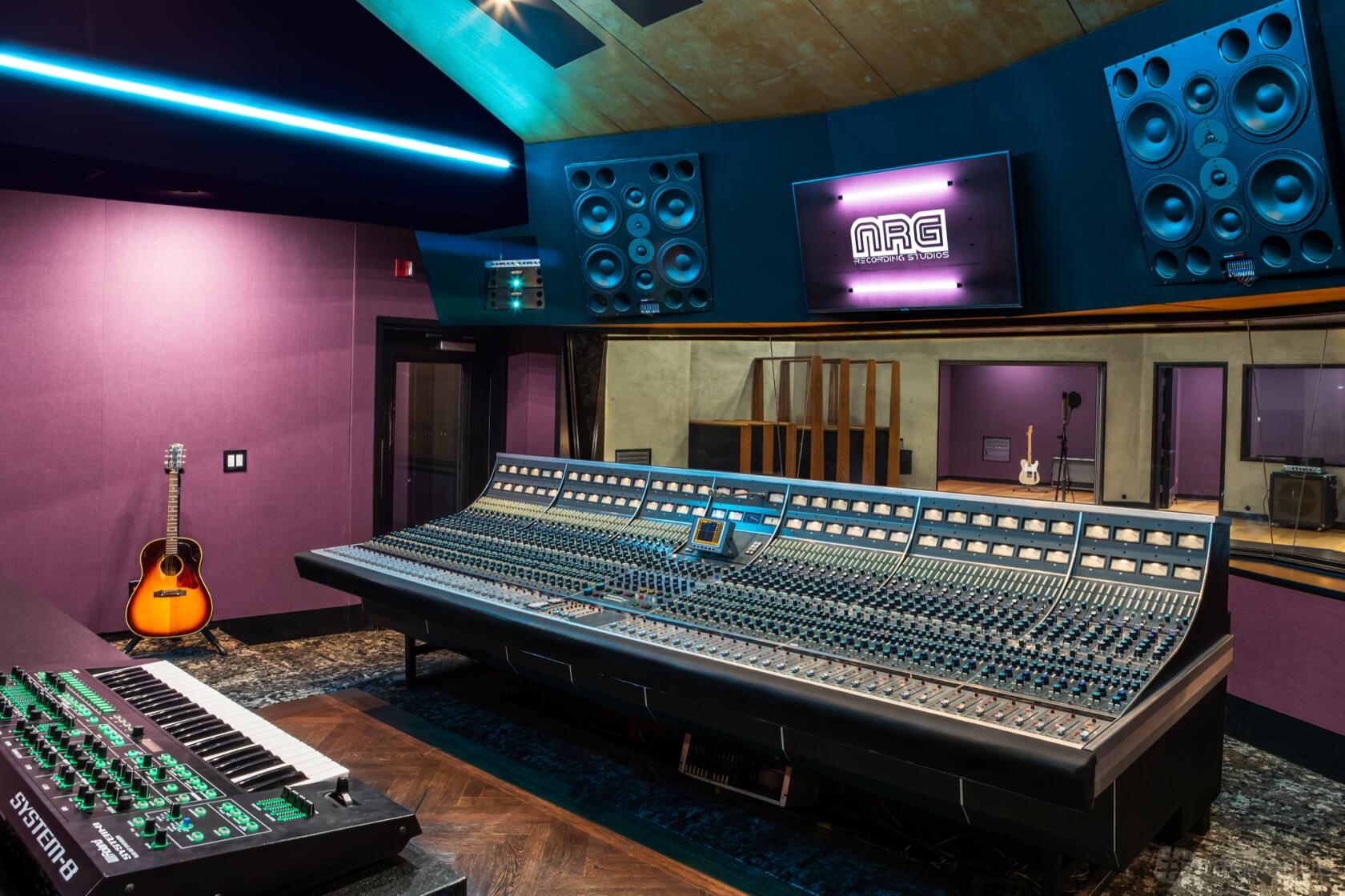 A recording studio with a large mixing console, wall-mounted speakers, and purple acoustic panels at NRG Recording Studios.