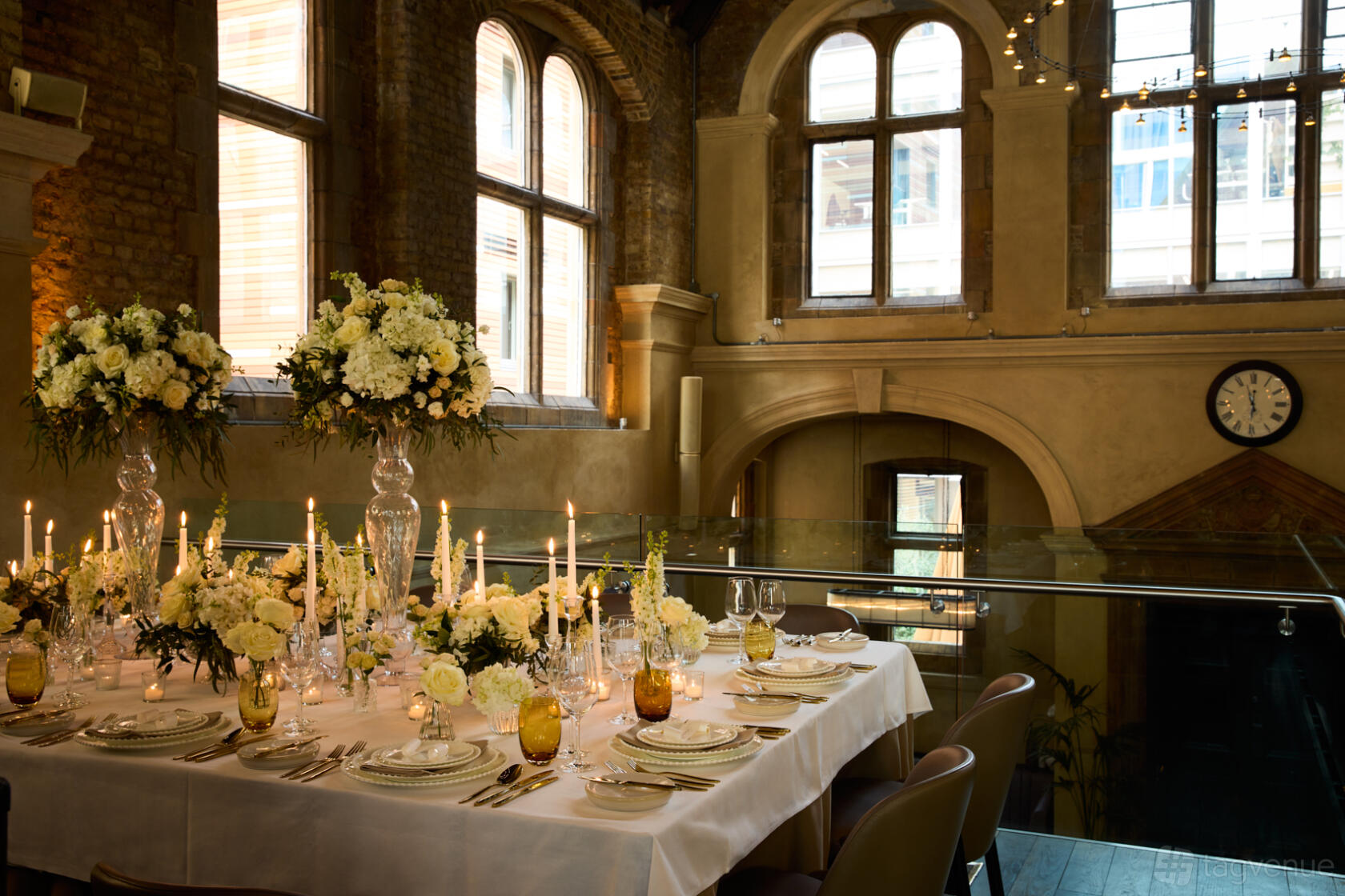 A dining room with tall arched windows, exposed brick walls, and a long table set with candles and floral centerpieces at Galvin La Chapelle.