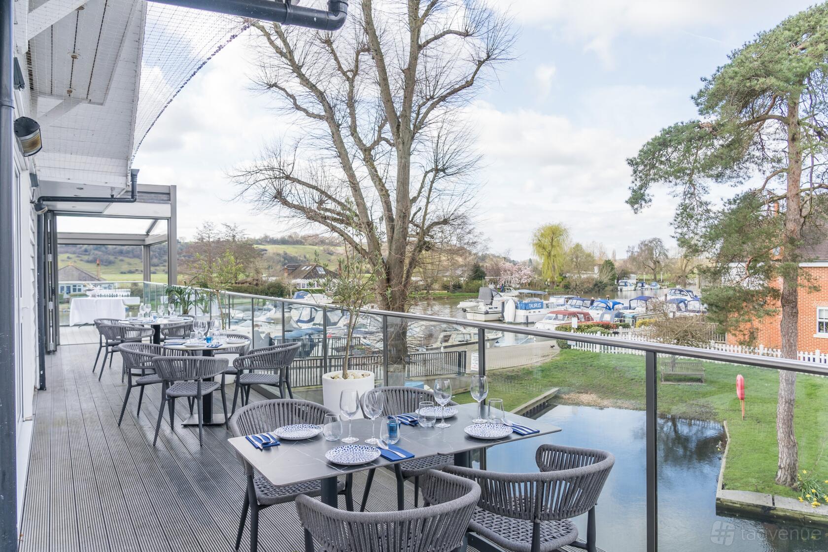 A restaurant terrace with glass railings, outdoor dining tables, and river views at El Cerdo Bourne End.