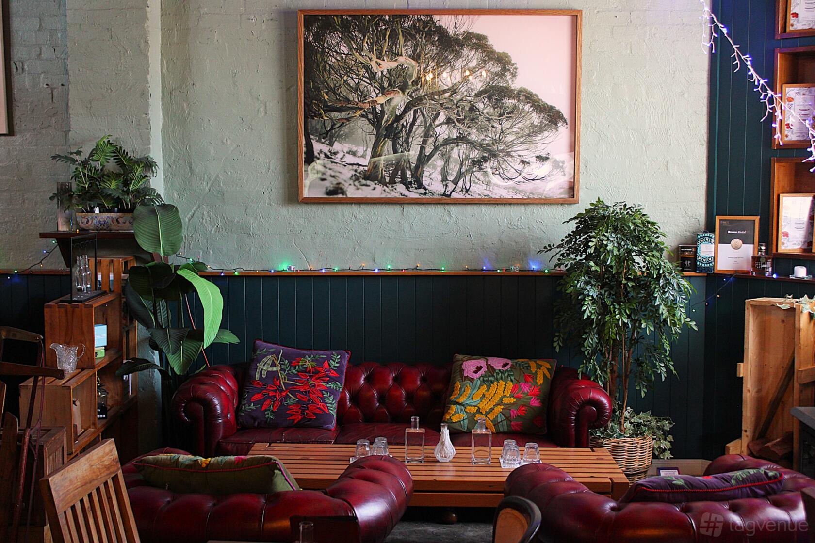 A bar lounge with red leather sofas, wooden tables, potted plants, and a large framed artwork at Goodradigbee Distillers.
