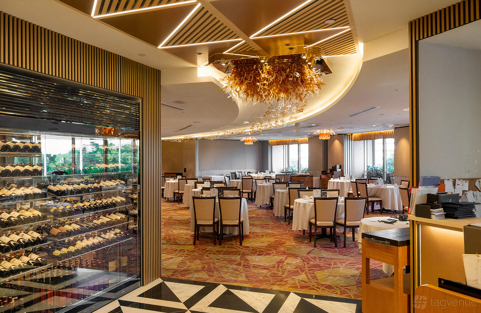 A restaurant dining room with round tables, cream chairs, chandeliers, and a glass wine display at Asia Grand Restaurant.