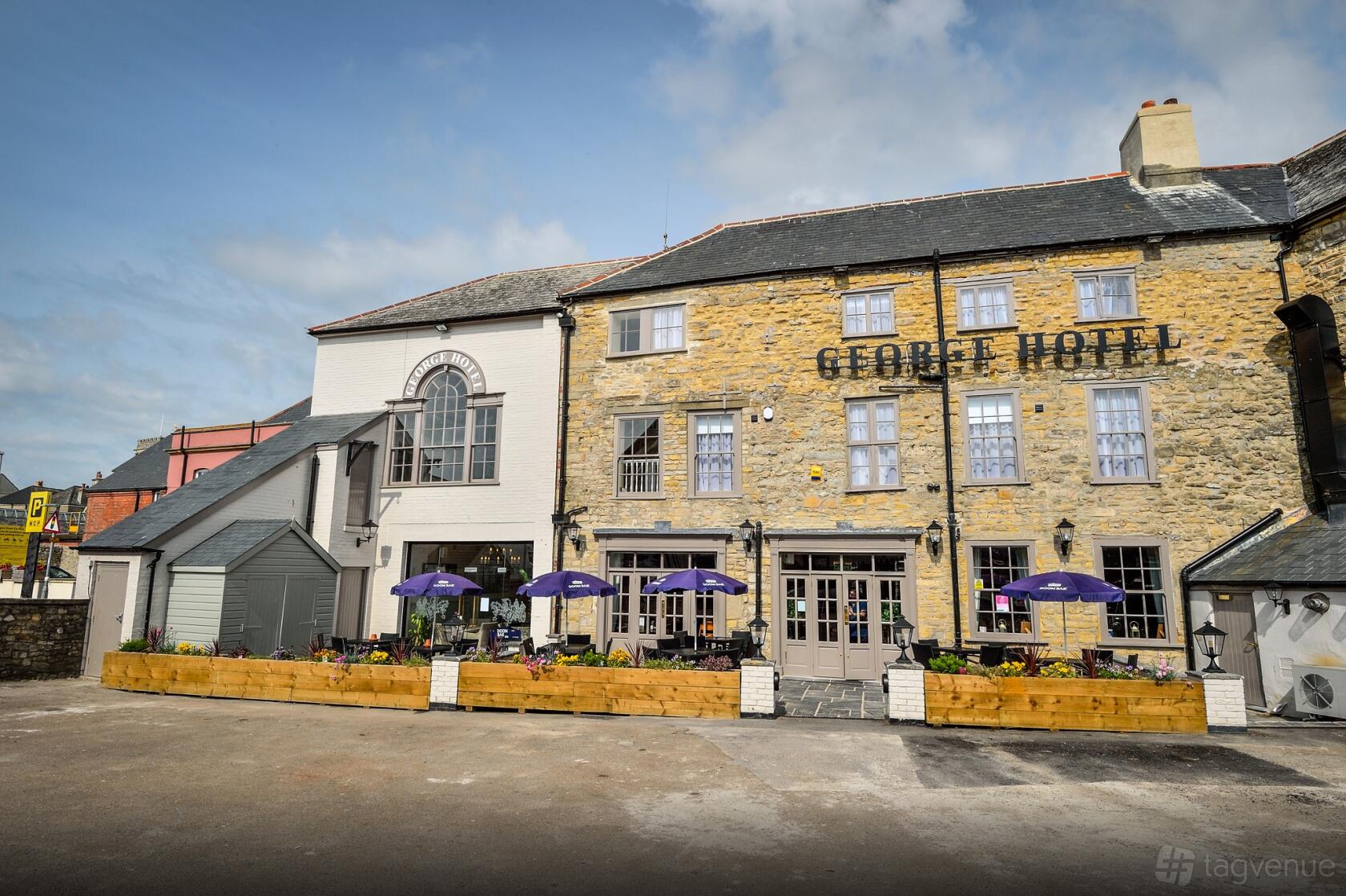 A historic stone hotel exterior with large windows, outdoor seating under purple umbrellas at The George Hotel.