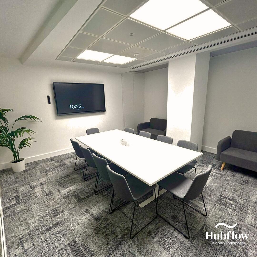 A meeting room with a long white table, gray chairs, wall-mounted screen, and carpeting at Hubflow Ltd