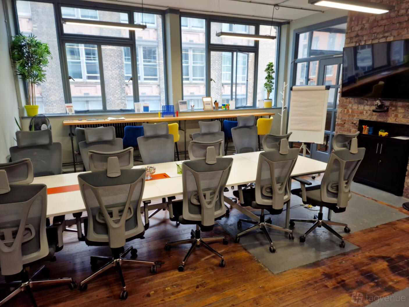 A meeting room with ergonomic chairs, long white tables, large windows, and exposed brick at Prime Meeting/Workspace at Cotton Exchange.
