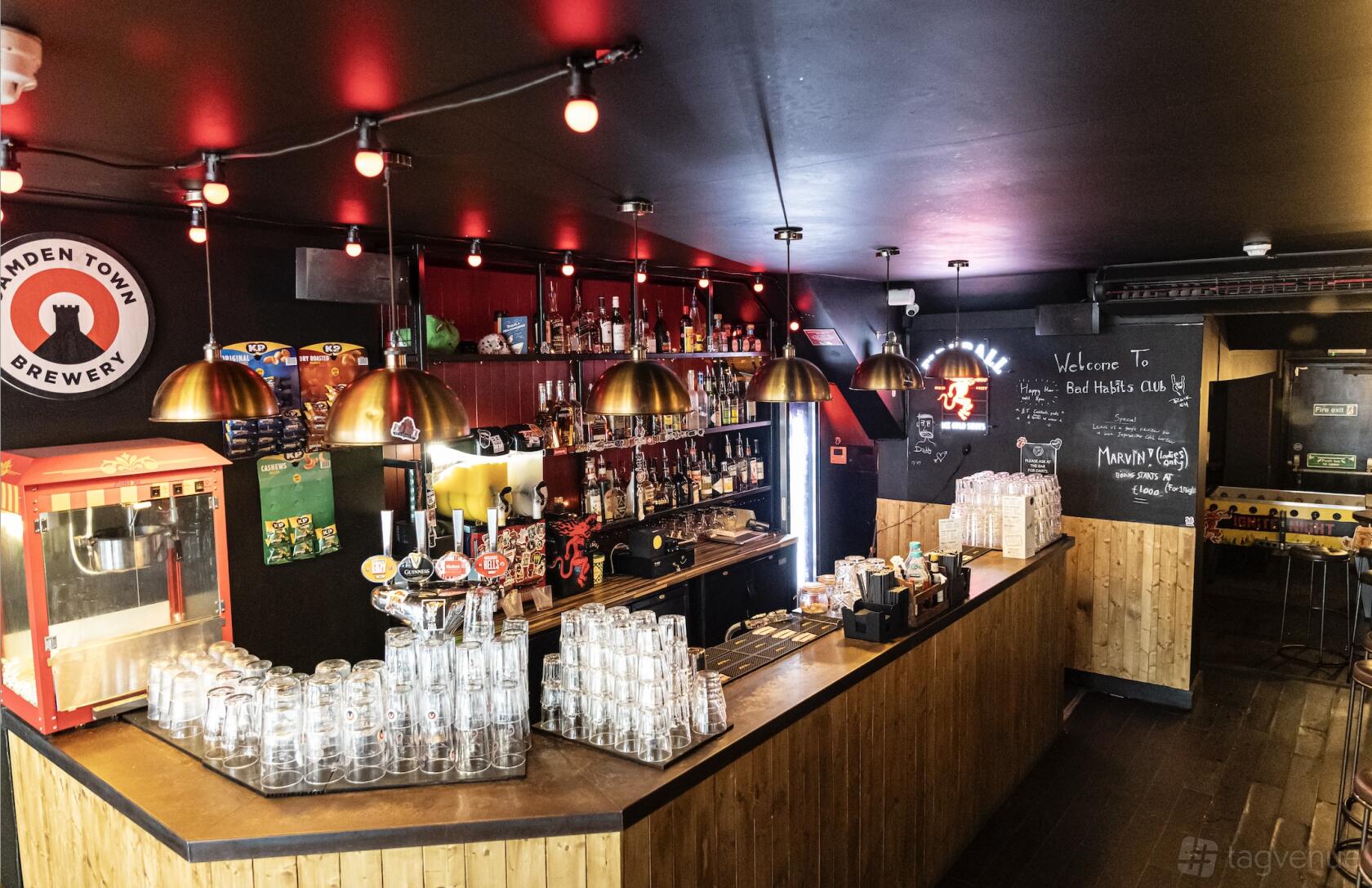 A bar with a wooden counter, hanging pendant lights, glassware, and shelves of spirits at Bad Habits Club.