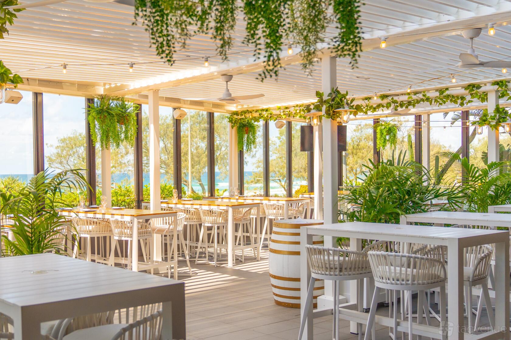 A terrace at a restaurant with white tables, hanging greenery, and ocean views at Coast Beach Bar & Kitchen.