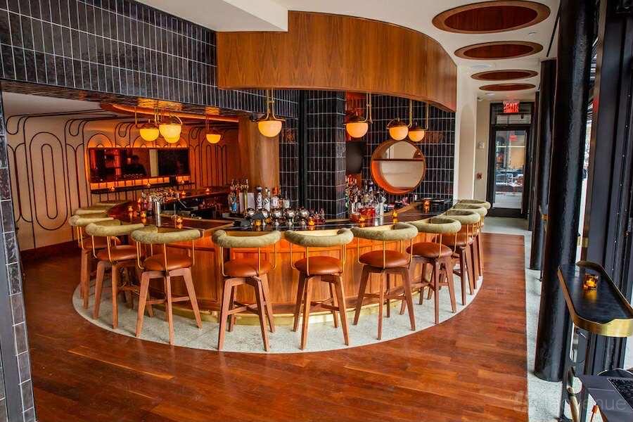 A curved wood bar with green velvet stools, globe pendant lights, and floor-to-ceiling windows at The Merchant Hotel.