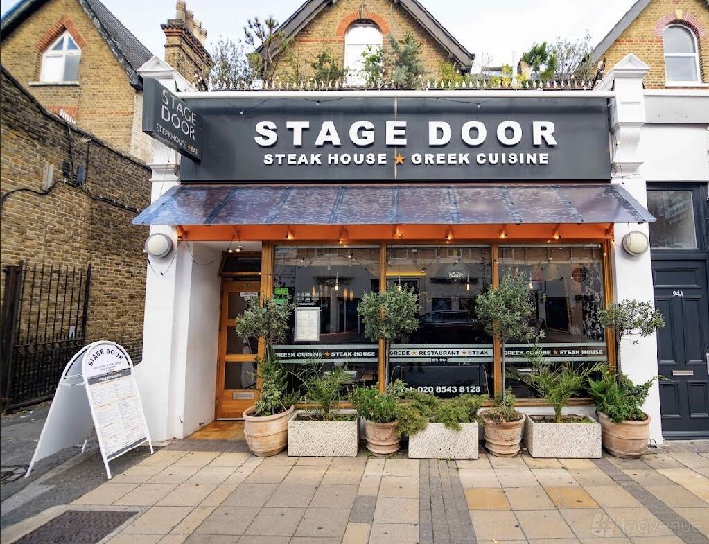 A restaurant with a glass storefront, orange awning, and potted plants at Stage Door Steakhouse Restaurant.