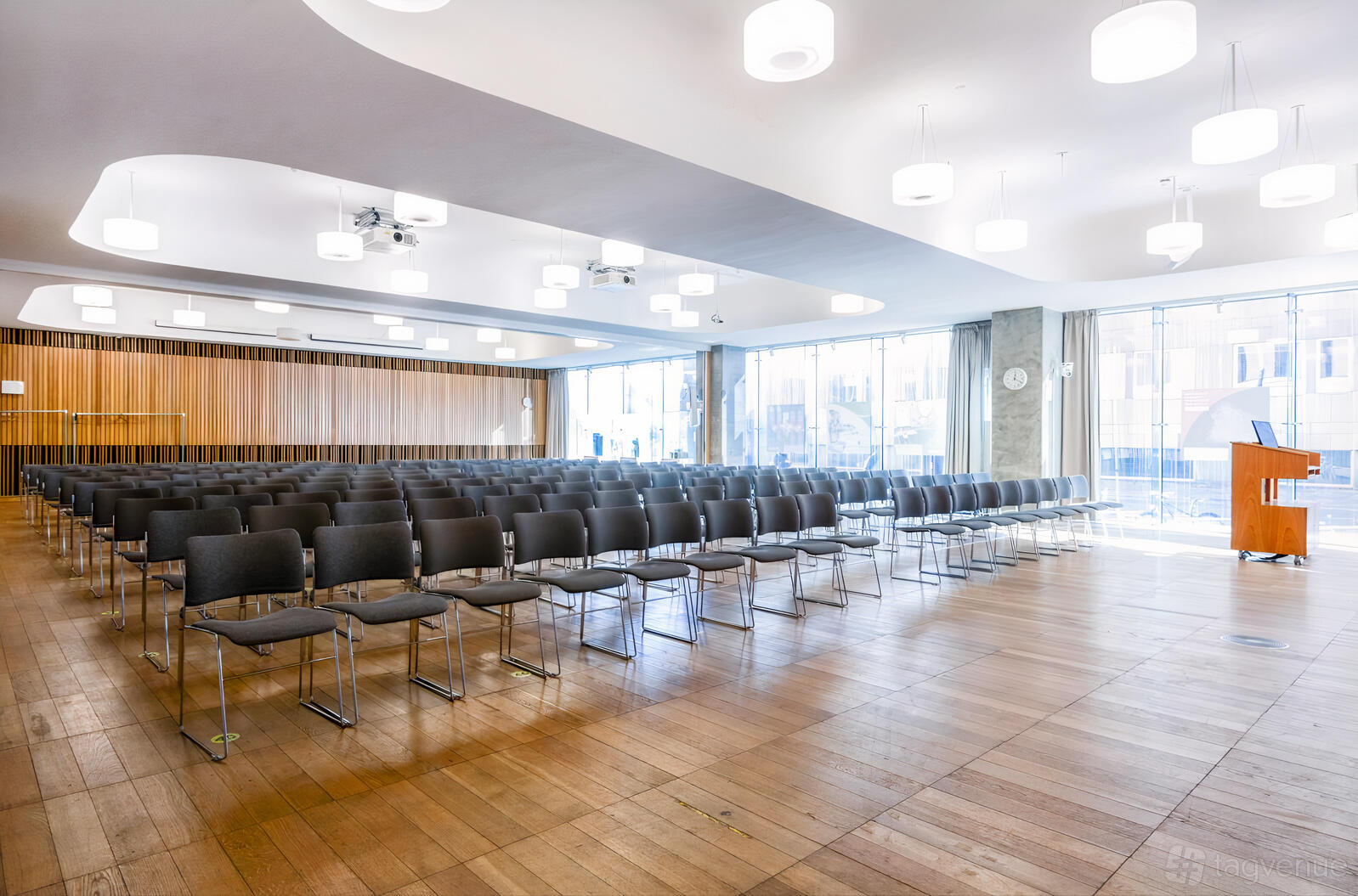 A university hall with rows of black chairs, wood flooring, large windows, and circular ceiling lights at Informatics Forum.