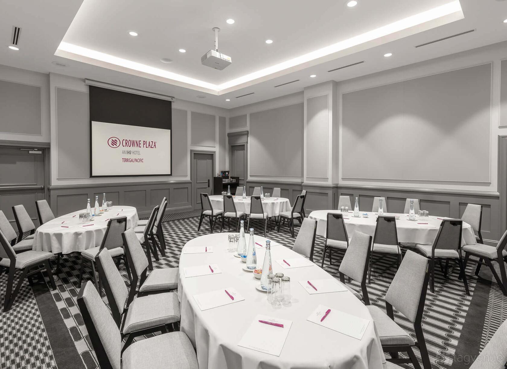 An event space with round tables, white linens, notepads, and a projector screen at Crowne Plaza Terrigal Pacific.