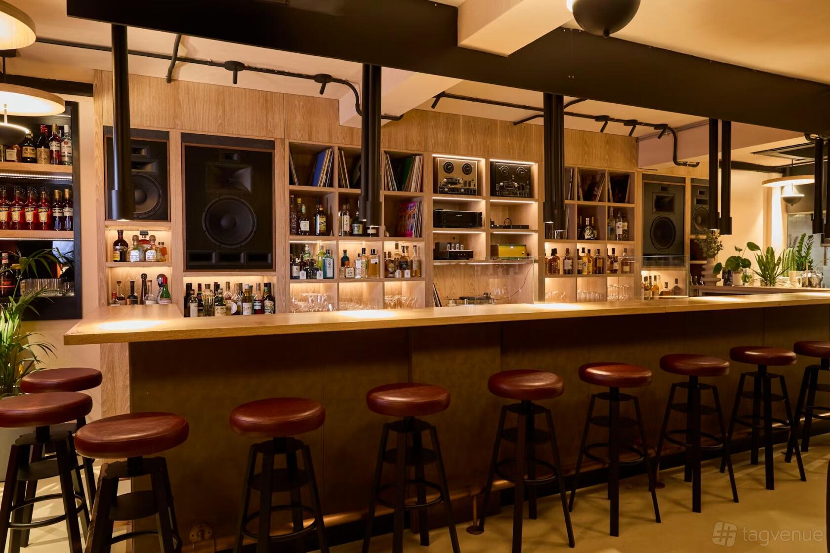 A bar with a long wooden counter, high stools, built-in shelves with bottles, and a sound system at Under The Counter.