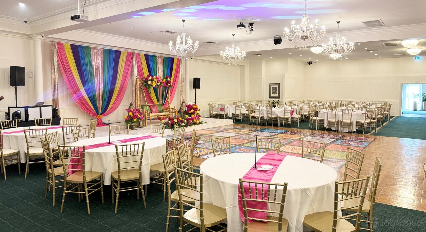 An event venue with round tables, gold chairs, chandeliers, and a stage with colorful drapes at Allure by Delhi Heights Function Centre.