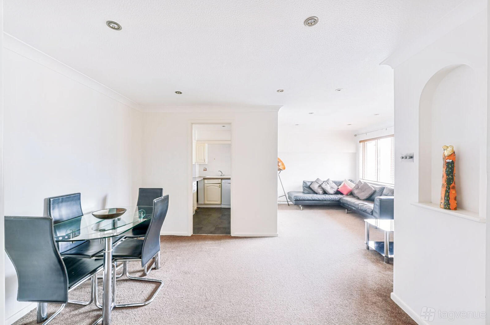 An apartment with a glass dining table, black chairs, neutral carpeting, and a lounge area with a sectional sofa at Cherry Court.
