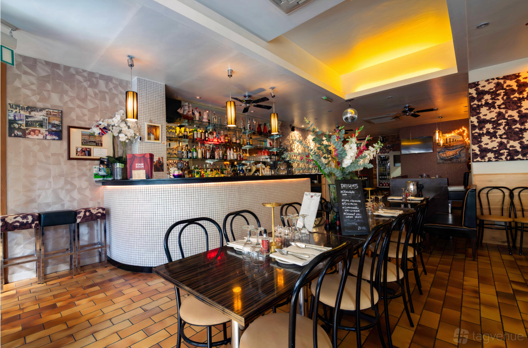 A restaurant with a tiled bar, wall-mounted shelves of liquor, wood tables, and decorative lighting at Stage Door Steakhouse Restaurant.