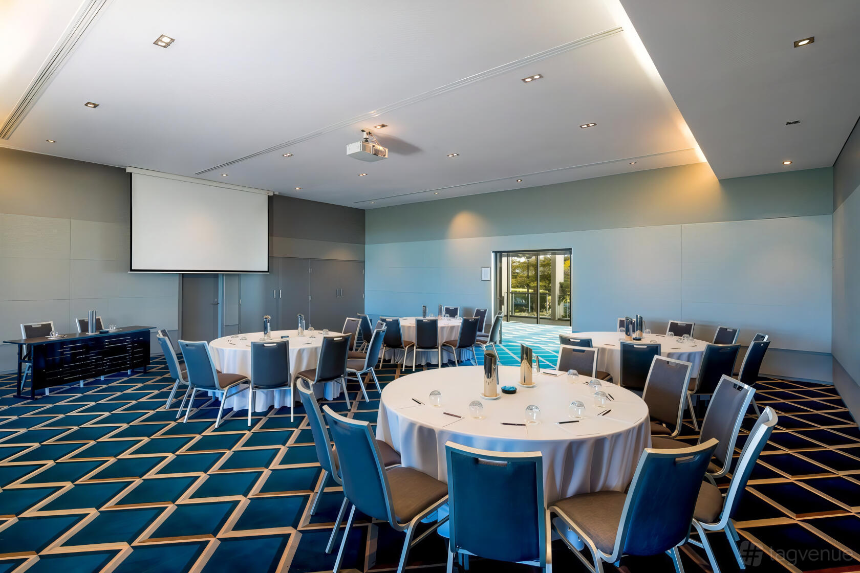 A function room with round tables, blue patterned carpet, and built-in projector at Pullman Magenta Shores Resort.