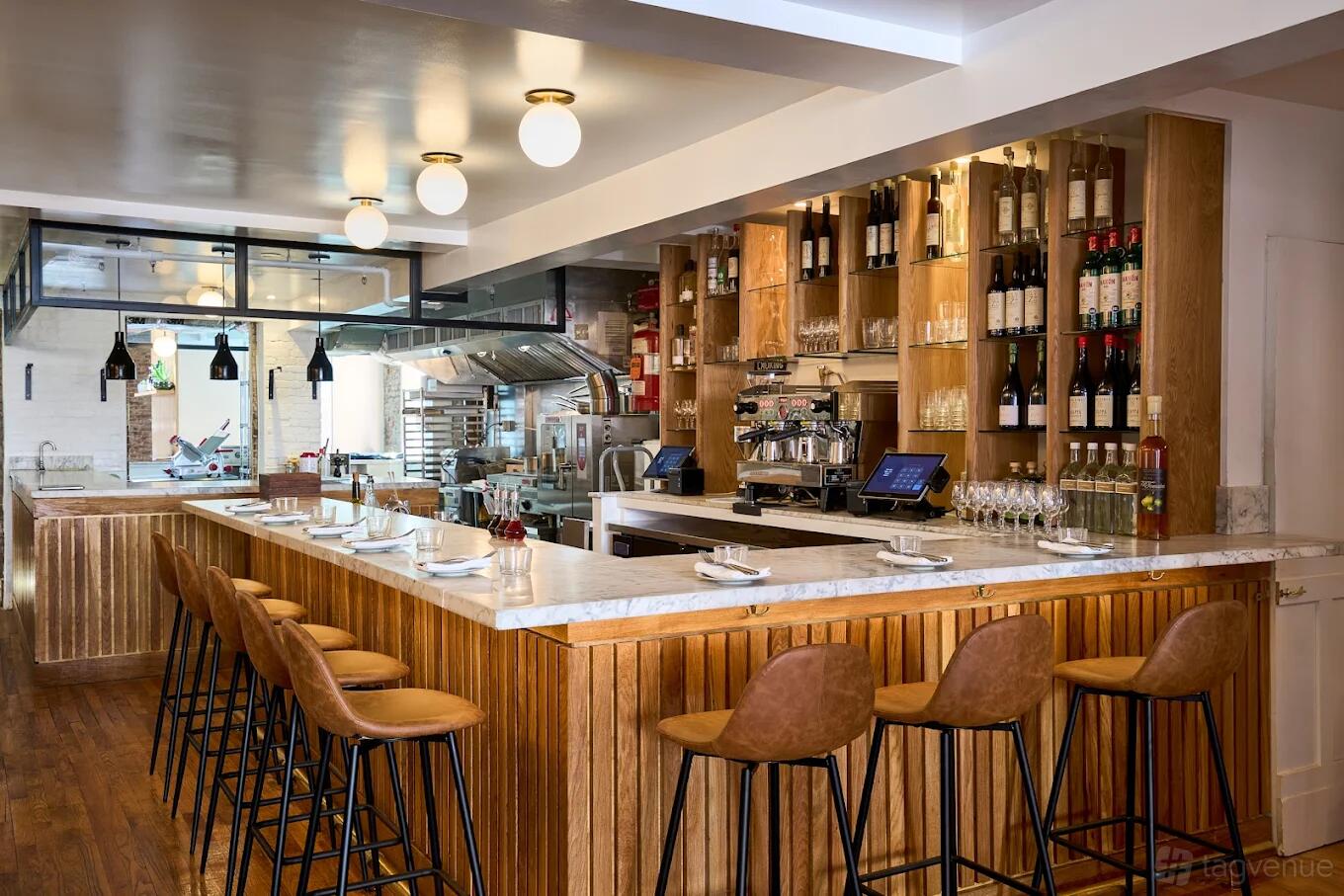 A restaurant with a marble-topped bar, wooden shelving displaying wine bottles, and tan barstools at Place des Fêtes.