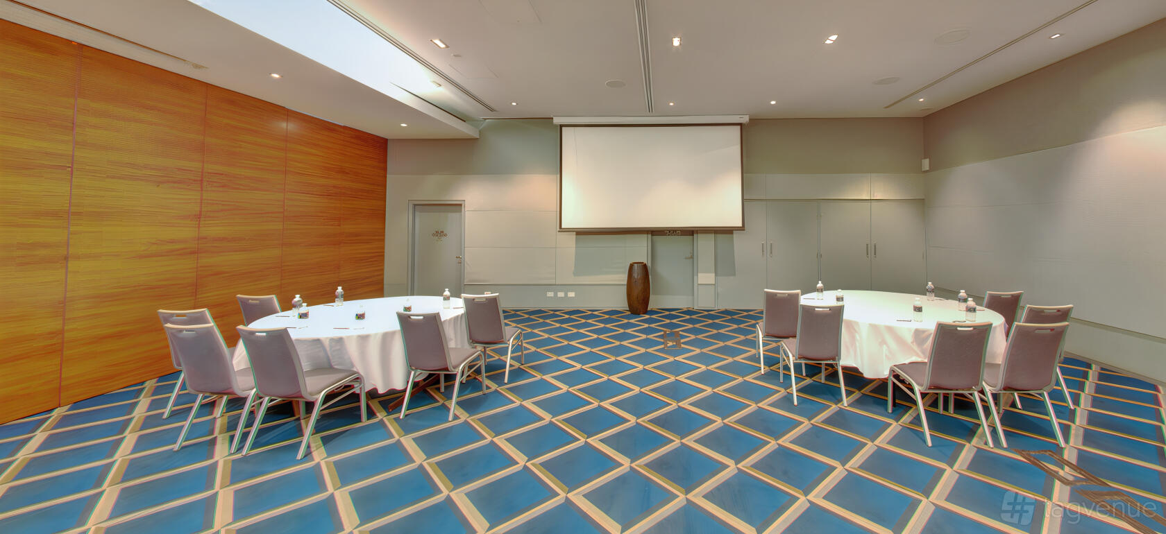 An auditorium with round tables, grey chairs, a projector screen, and patterned blue flooring at Pullman Magenta Shores Resort.