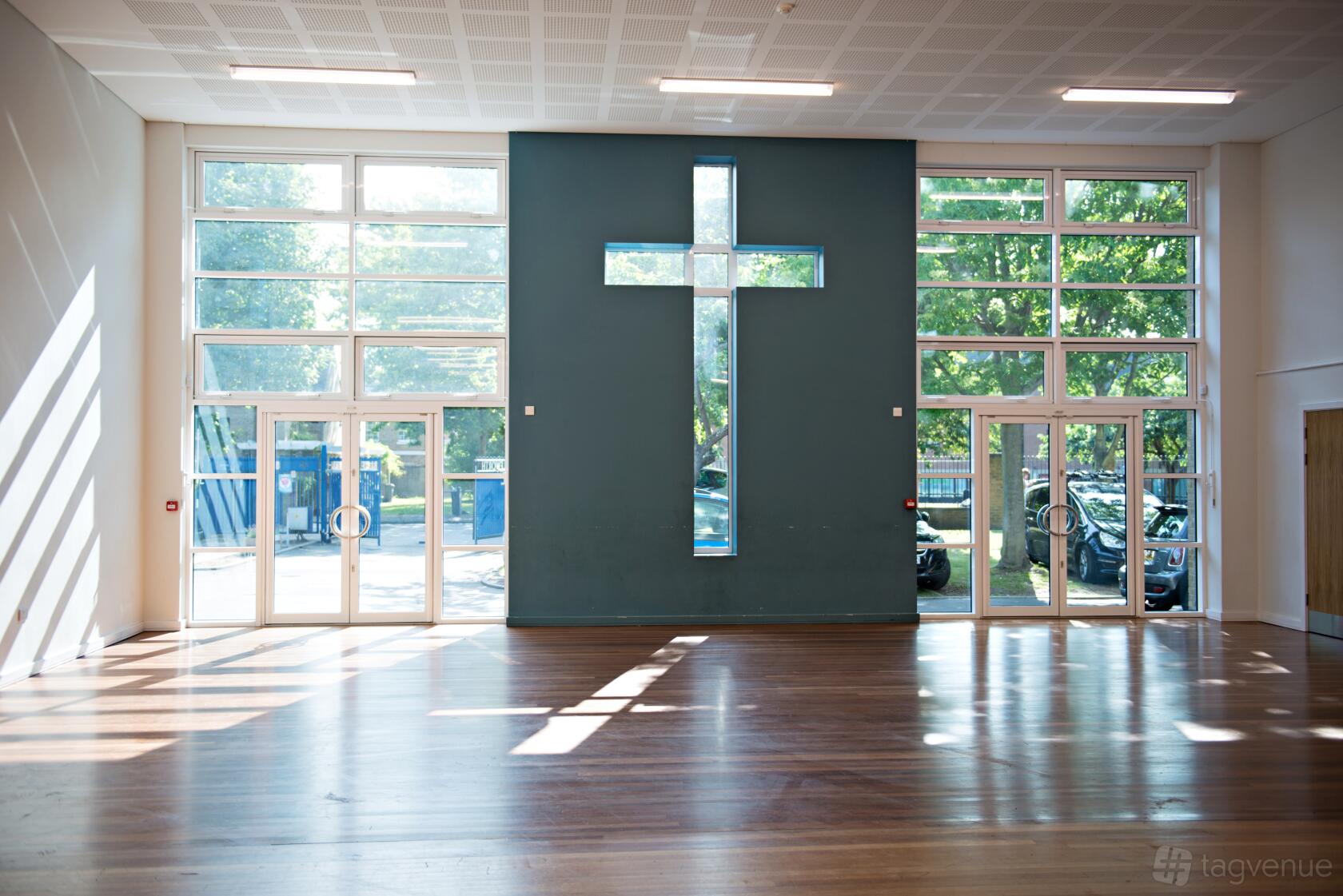 An event hall with large floor-to-ceiling windows, wooden flooring, and a cross-shaped window at The Blue school hall.