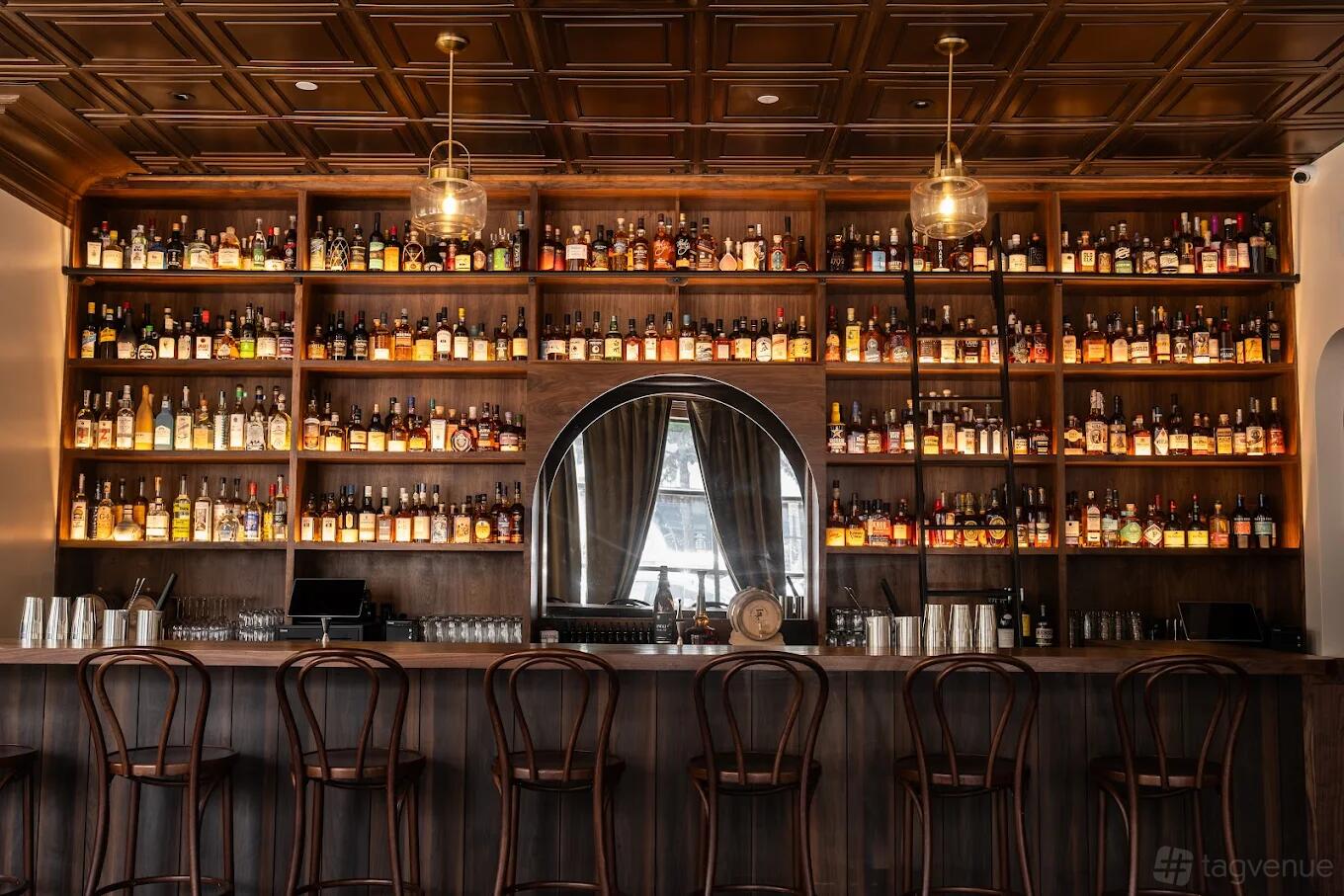 A whiskey bar with a long wooden counter, high stools, and a backlit wall of liquor bottles at Foolproof Whiskey Bar.