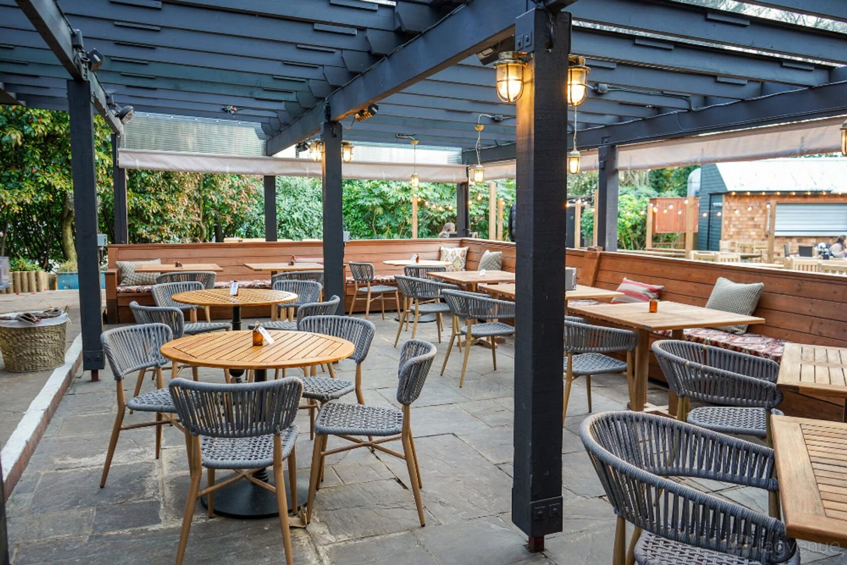 An outdoor pub patio with wooden round tables, woven chairs, overhead pergola, and benches at The Leather Bottle.