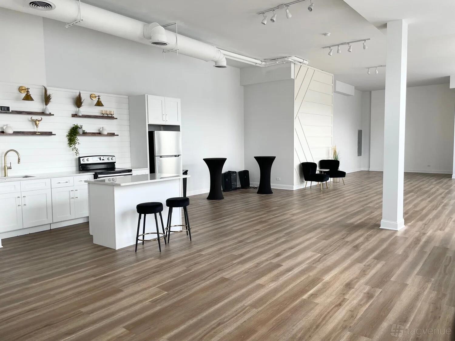 An event venue with light wood floors, white walls, a kitchenette, and scattered black chairs at Gallery 86.