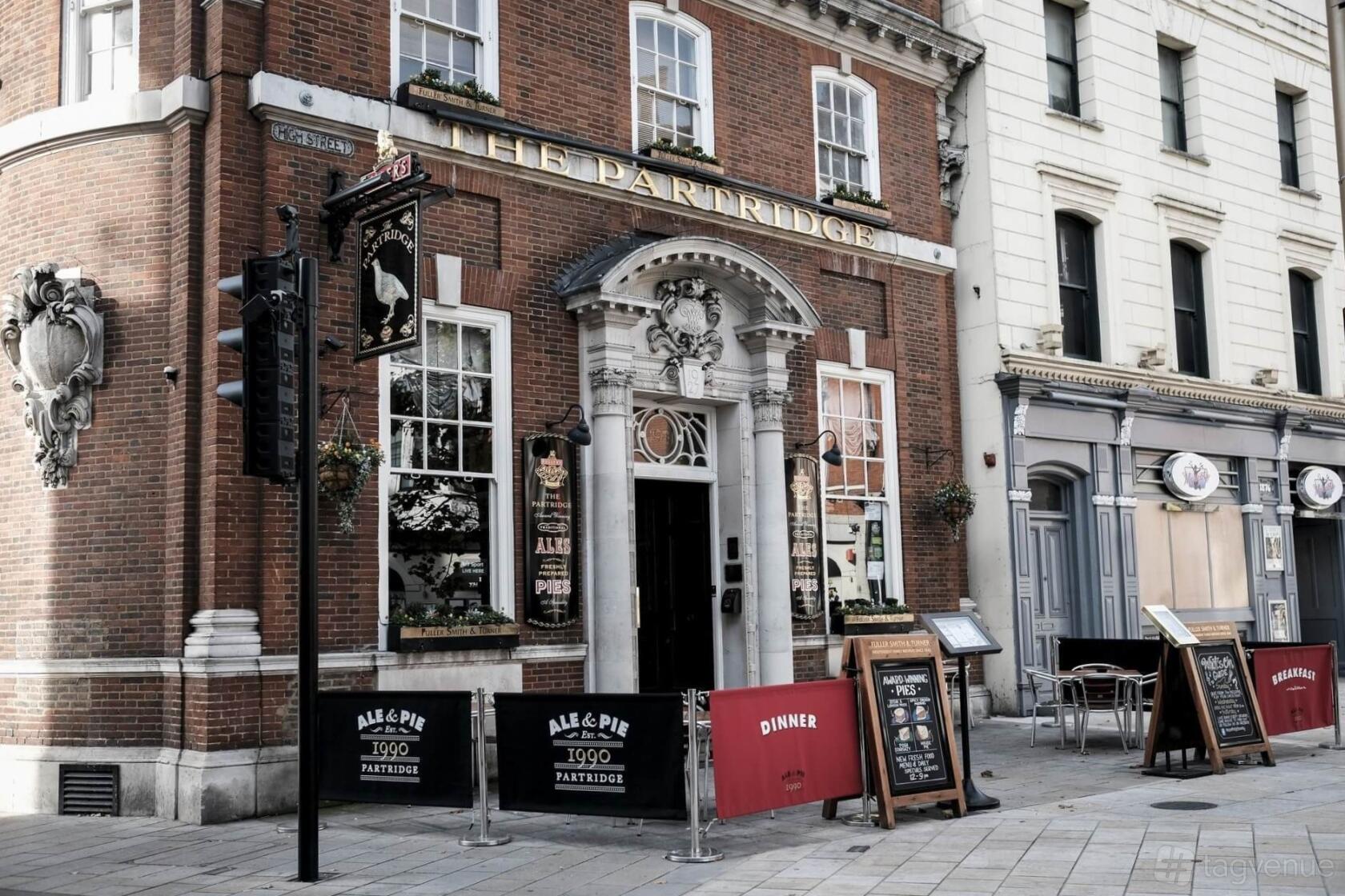 A pub with a red brick facade, arched entrance, hanging flower baskets, and outdoor seating at The Partridge, Bromley.
