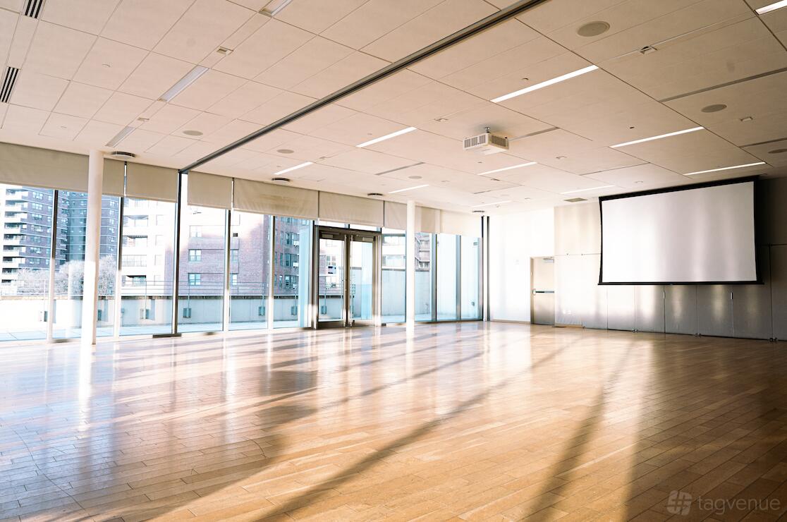 An event space on a rooftop with floor-to-ceiling windows, hardwood floors, and a ceiling-mounted projector screen at The Rooftop @ Manny Cantor Center.