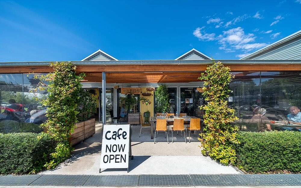 A café with outdoor seating, glass walls, greenery, and a 'cafe now open' sign at Fork N Flower.