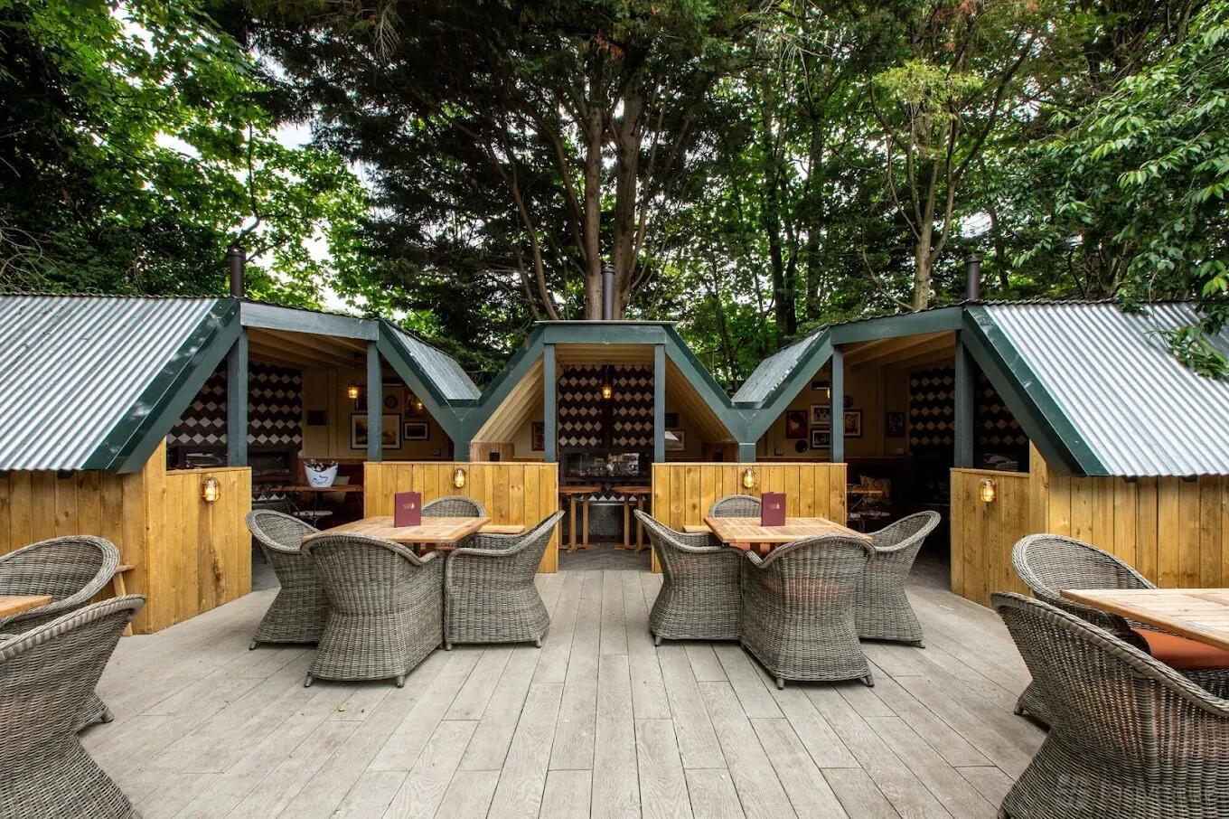 An outdoor pub space with wicker chairs, wooden decking, and individual hut seating at The Leather Bottle.