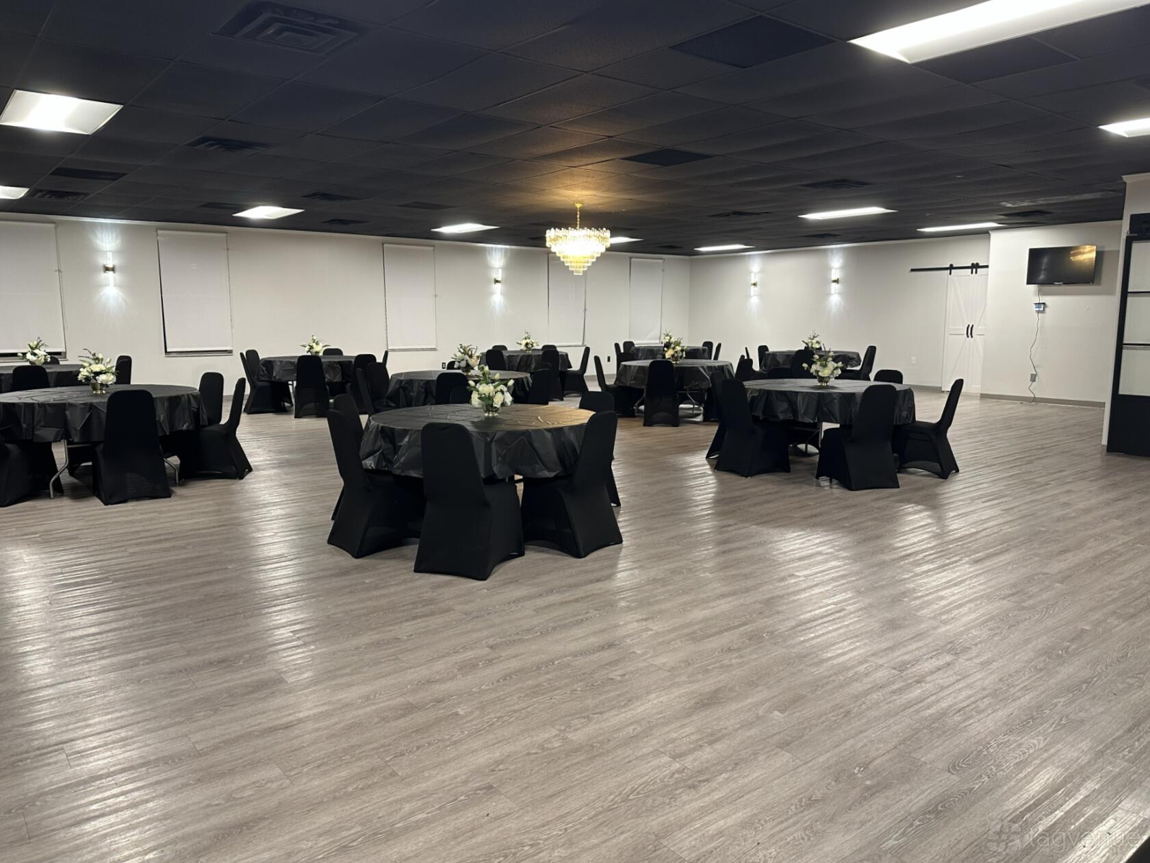 A ballroom with black round tables, matching covered chairs, wood flooring, and a central chandelier at Miller Legacy Event Center.