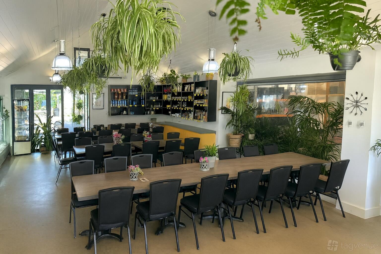 An event space in a cafe with long wooden tables, black chairs, hanging plants, and large windows at Fork N Flower.