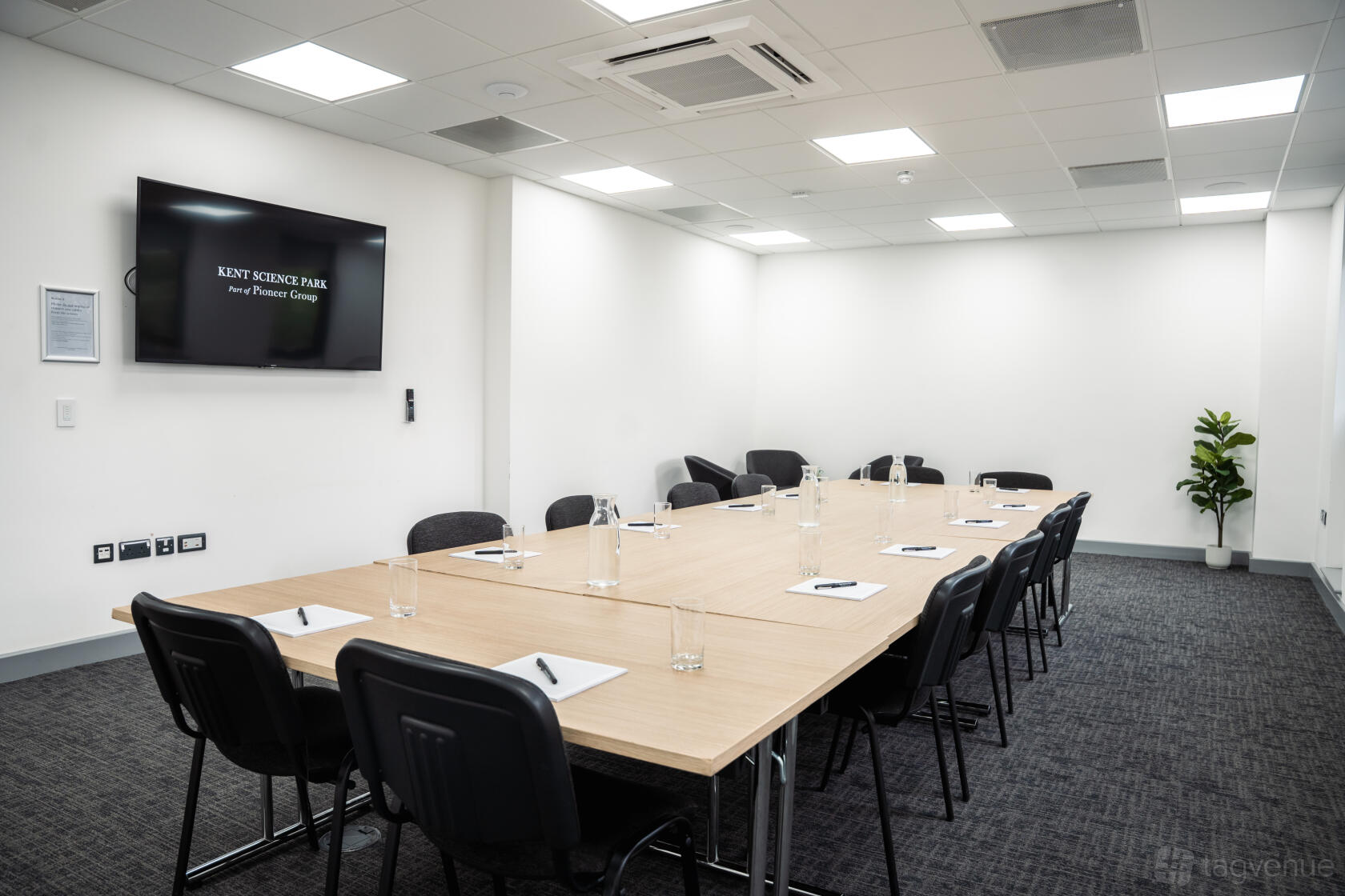 A meeting room with a U-shaped table setup, black chairs, wall-mounted screen, and notepads at Kent Science Park.