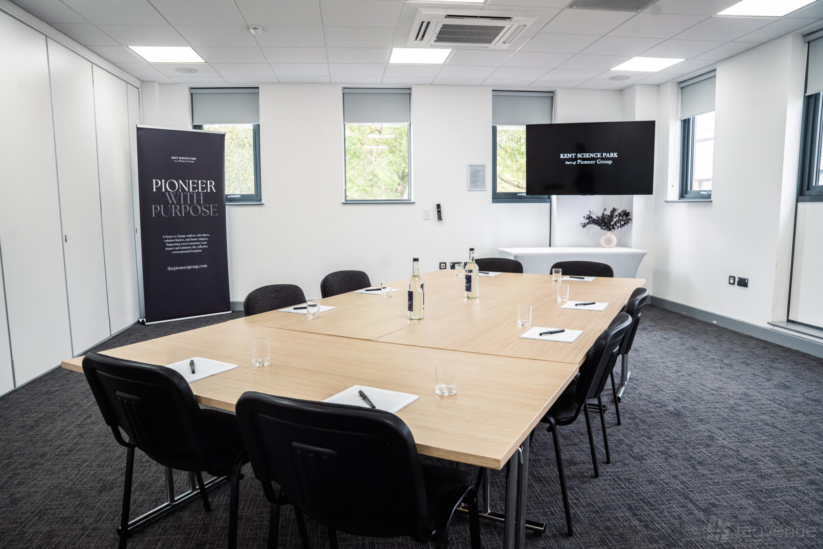 A meeting room with a U-shaped wooden table, black chairs, notepads, and a wall-mounted screen at Kent Science Park.
